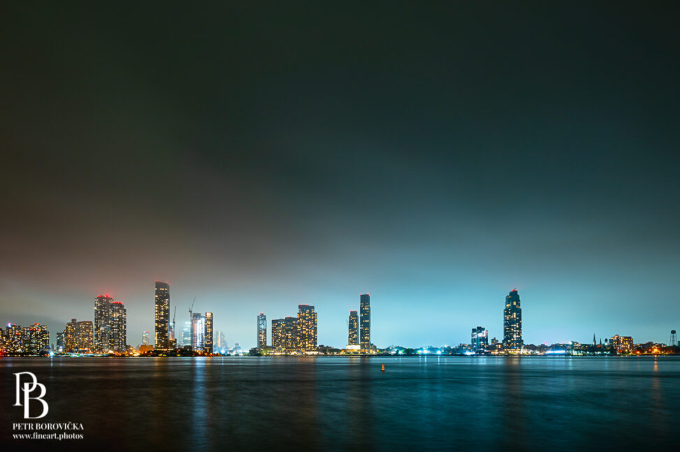Long exposure view of the New Jersey skyline as seen from New York, featuring smooth water of the Hudson River and stark architectural silhouettes.