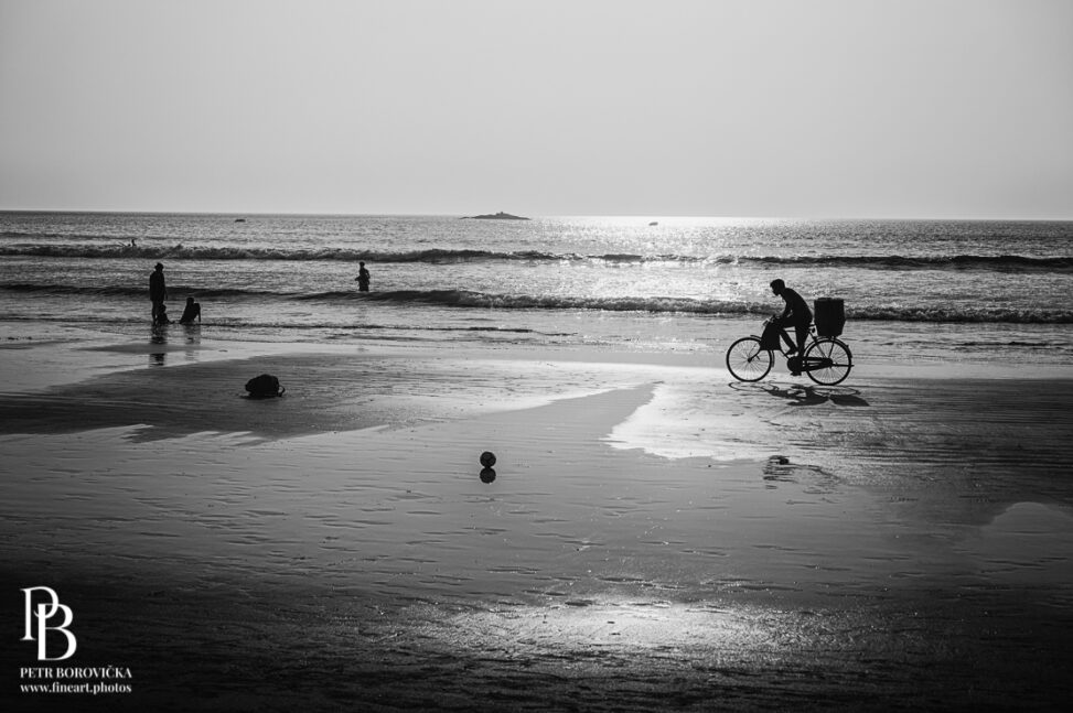 High contrast black and white view of silhouetted people walking across a beach in Gokarna, India, casting long shadows in the late afternoon sun.