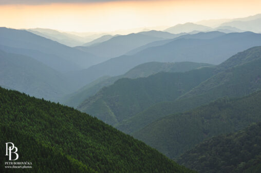 Layered green mountain ridges in Japan receding into a hazy sunset sky.