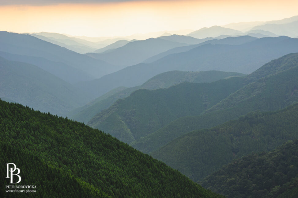 Layered green mountain ridges in Japan receding into a hazy sunset sky.