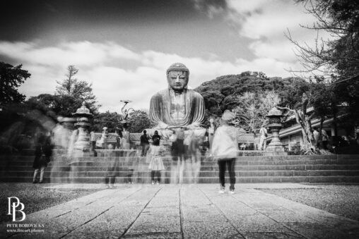 High contrast black and white long exposure of the monumental Kamakura Daibutsu, with the blurred silhouettes of passing figures moving in the foreground.